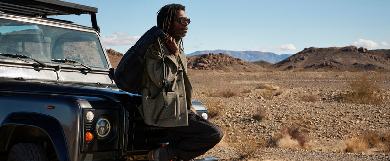 A man is shown leaning against the hood of his classic SUV, wearing wool jacket that is layered.