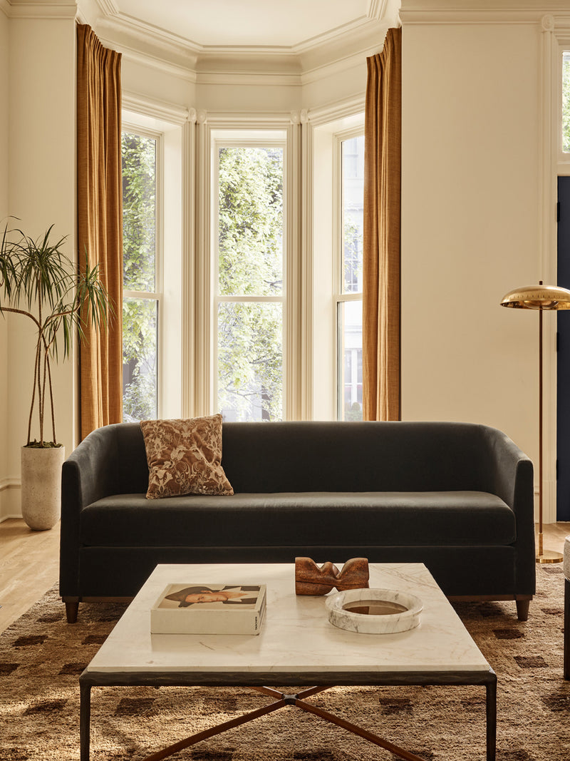 Dark blue sofa in a living room. Behind it is a brass floor lamp, potted palm, tall windows with golden drapes. In front is a square marble and metal coffee table with book, wooden sculpture and round marble mirror tray. There is a large brown rug with darker brown squares on the floor.